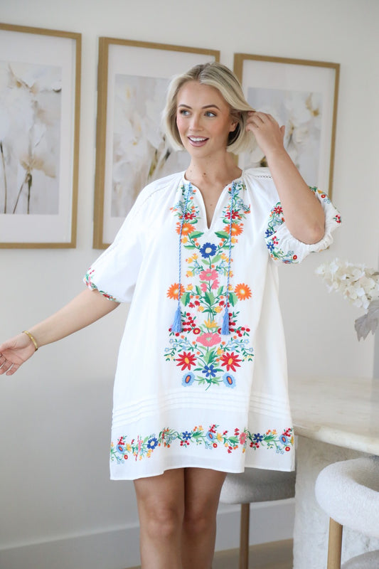 Woman wearing Solara Boheme Boho Bloom Mini Dress with colorful floral embroidery and tassel ties in bright indoor setting