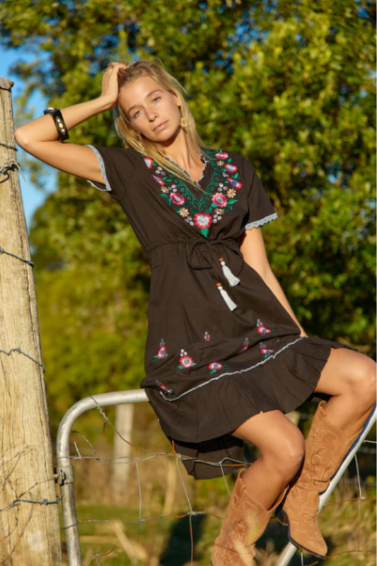 Woman wearing Solara Boheme Casey Mini Dress with floral embroidery sitting outdoors by a wooden fence in sunlight