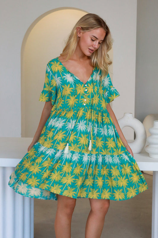 Woman wearing a teal Clara Mini Dress with yellow and white palm tree print, featuring buttons and tassel ties.