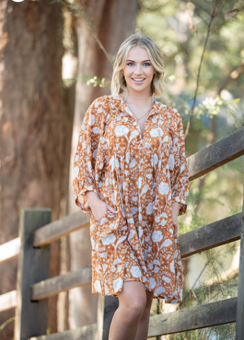 Smiling woman wearing Solara Boheme Fleur Mini Dress with earthy floral print and billowy sleeves outdoors by wooden fence