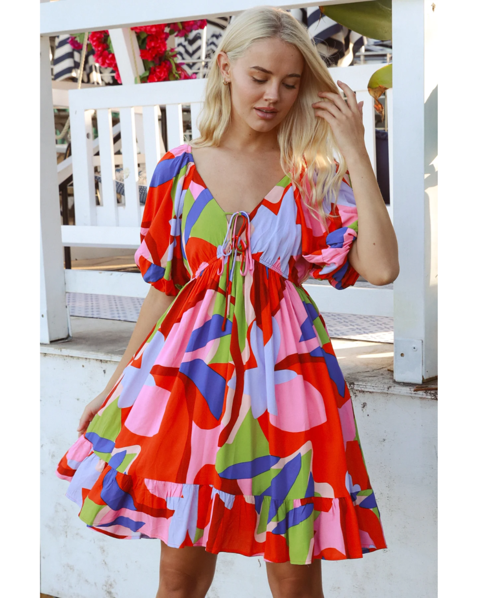 Woman wearing a colorful Alberta Mini Dress with puff sleeves and a tie-front detail, standing outdoors by white railing.