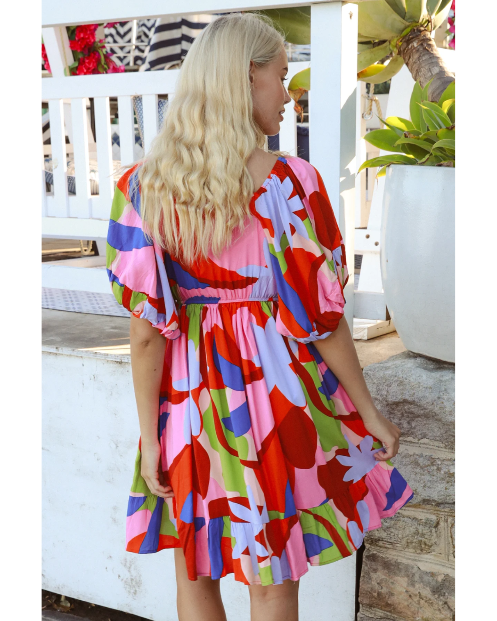 Woman wearing Avamia Alberta Mini Dress with colorful abstract print and puff sleeves, standing outdoors near plants and railing.