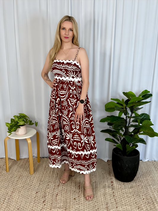 Woman wearing Alexa Midi Dress by Solara Boheme with bold brown and white print and Ric-Rac trim, standing indoors near plants