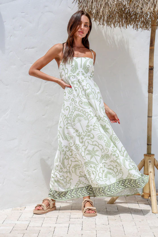 A woman stands outdoors in the Alora Maxi Dress—a long, white sleeveless dress with a pale green floral pattern in a rayon linen blend—by a textured white wall and straw parasol, sunlight casting soft shadows around her.