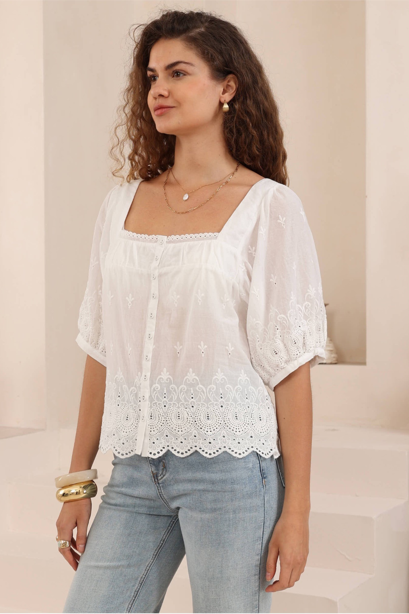 A woman wearing a white short-sleeve blouse with a square neckline, lace trim, and puff sleeves, paired with light blue jeans.