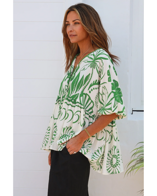 A woman with long brown hair stands sideways against a white wall, wearing the Annalise Blouse—crafted from breathable fabric with bold green floral patterns—and black pants.