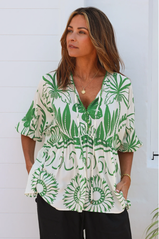 A woman with long brown hair wears the Annalise Blouse in a green and white floral print with breathable fabric, paired with black pants and gold jewelry, standing against a white wall and looking to the side.