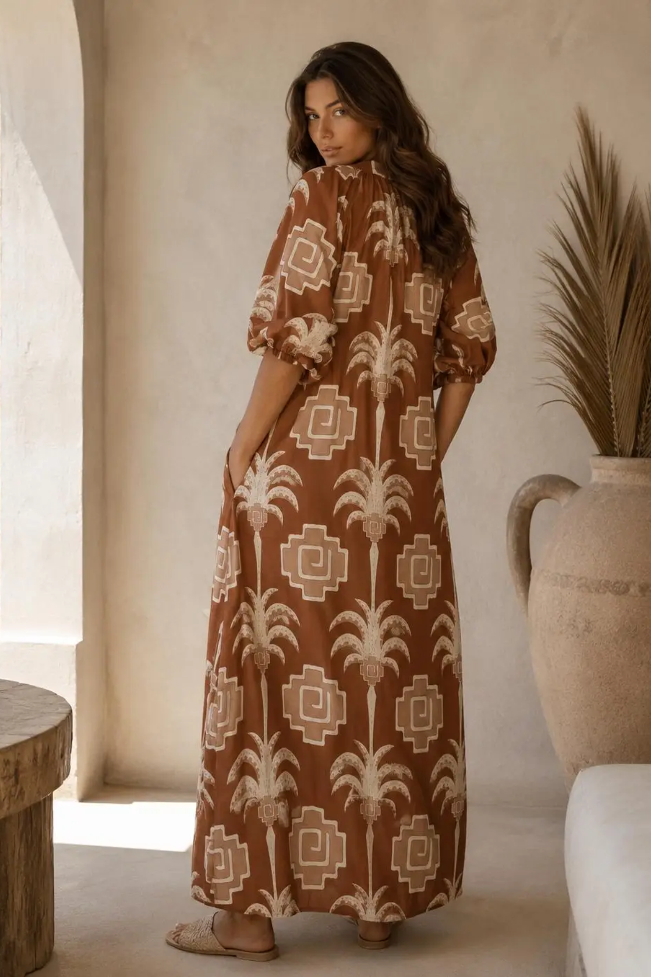 A woman wearing a brown and beige patterned maxi dress, standing in a warm-toned room with a large ceramic vase and dried palm leaves in the background.