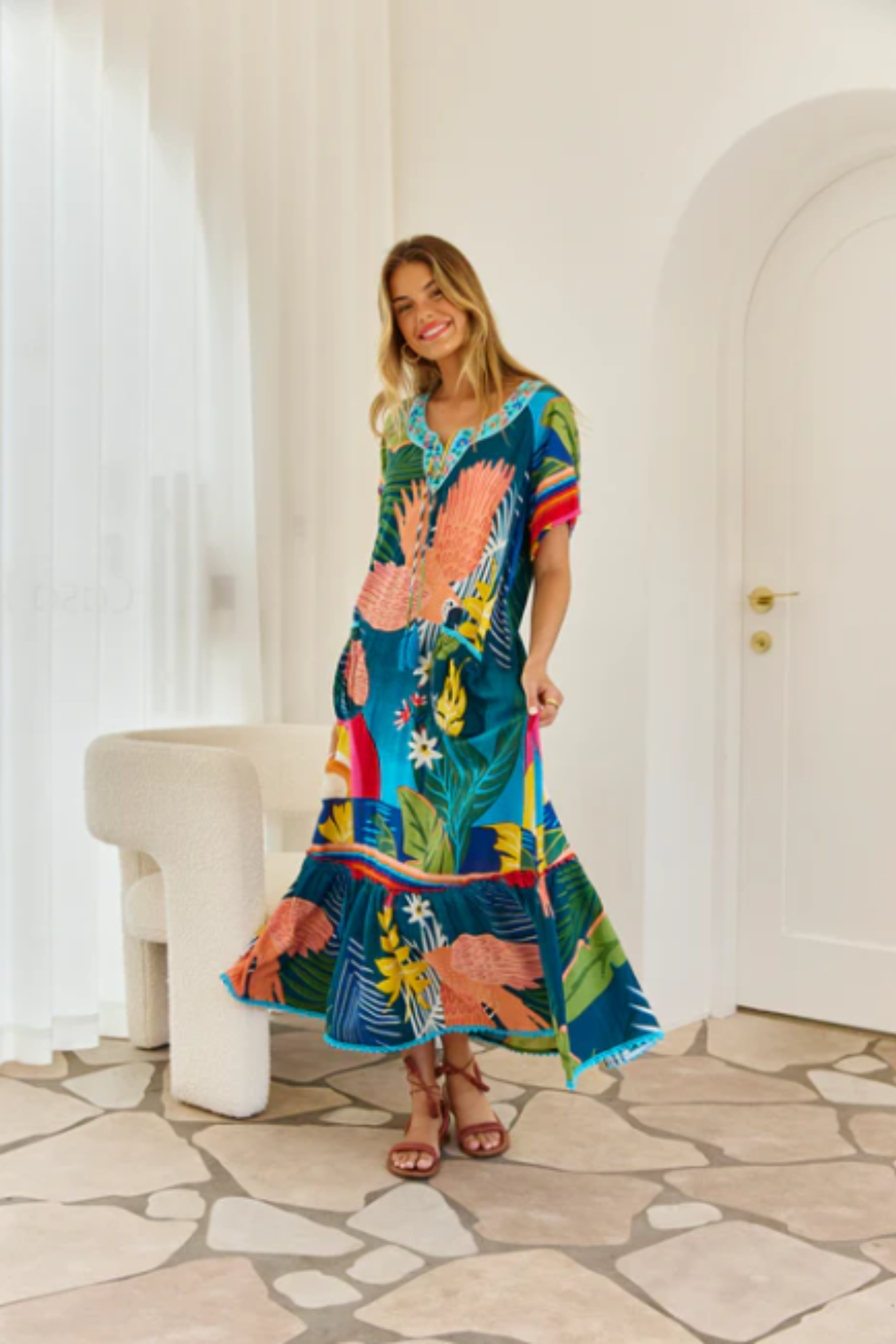 Woman wearing a vibrant Boojee Maxi Dress by Solara Boheme with tropical print and frilled hemline indoors.