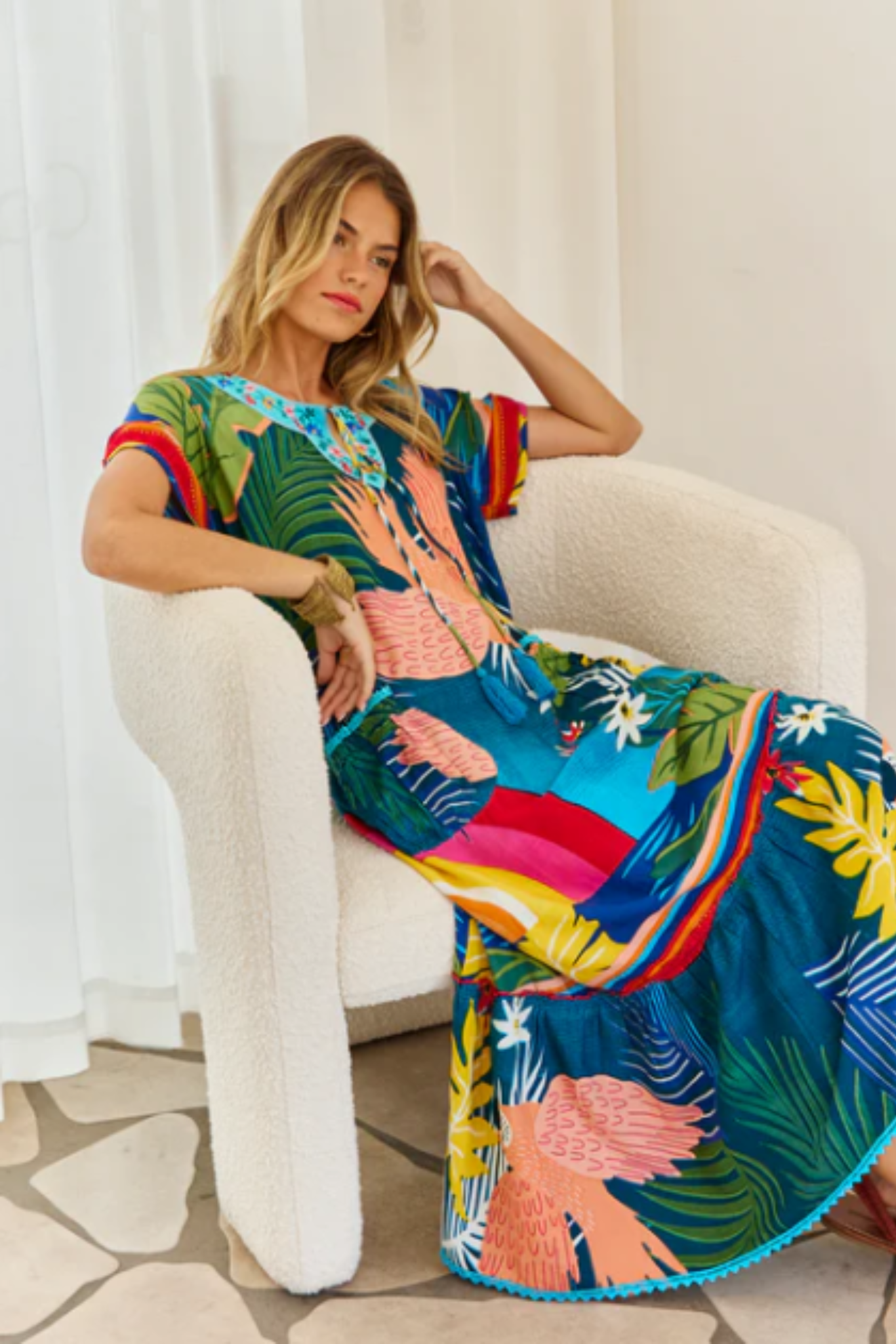 Woman seated in a white chair wearing Solara Boheme's vibrant Boojee Maxi Dress with tropical print and embroidered neckline