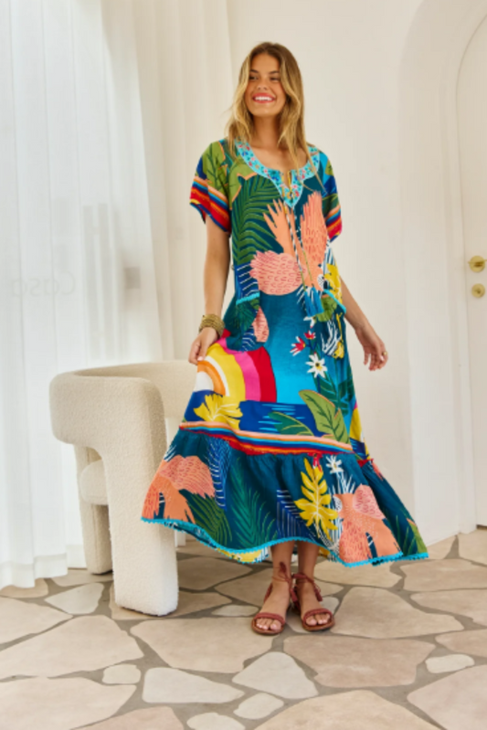 Woman wearing a vibrant Boojee Maxi Dress by Solara Boheme with tropical prints and tassel details indoors
