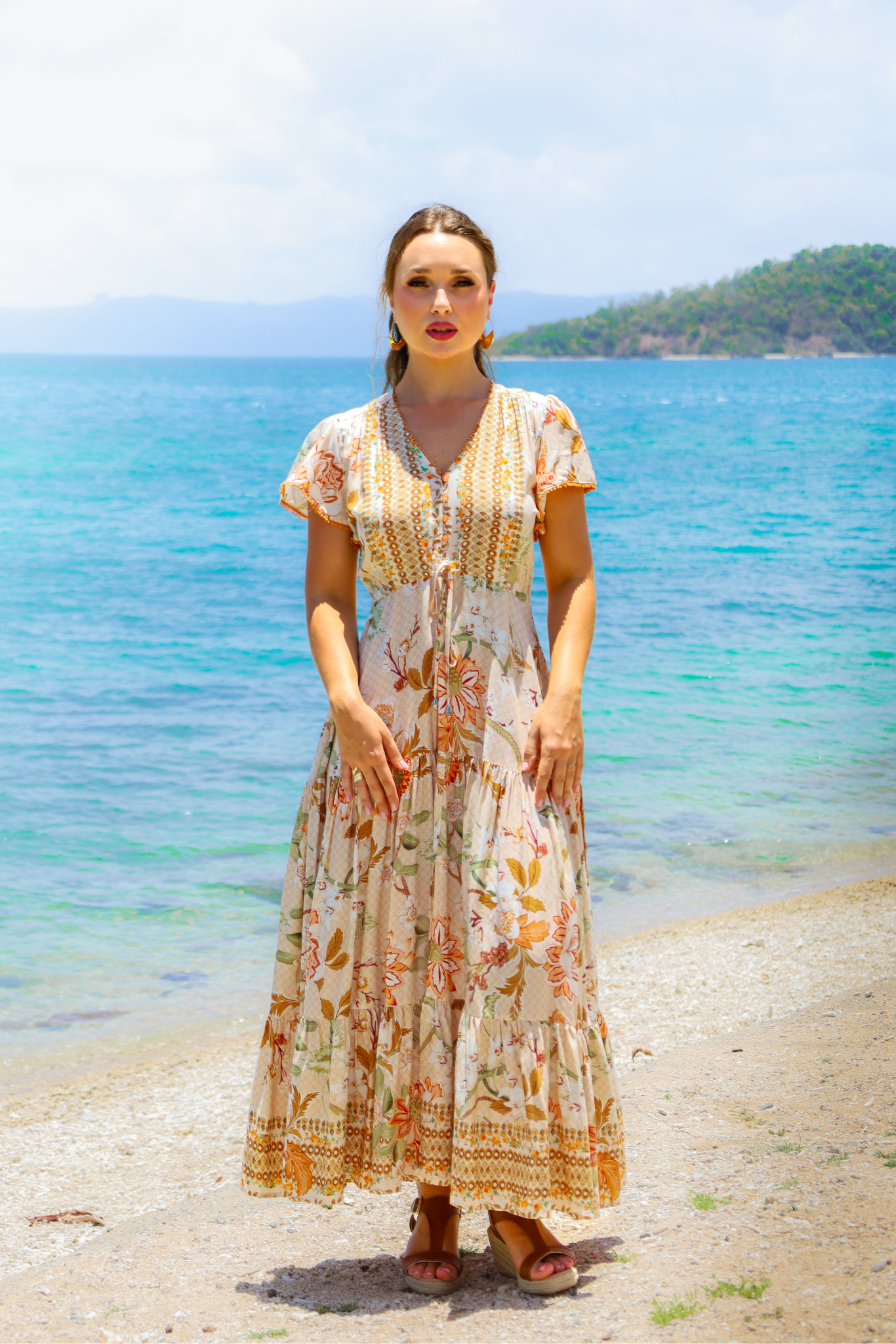 Woman in a floral dress standing on a beach with clear blue water and sky.