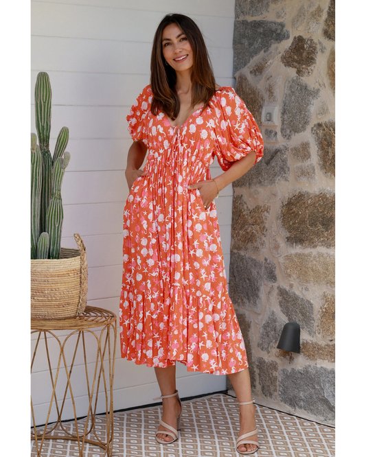 Woman wearing an orange Ava & Mia Chrysta V-Neck Midi Dress with white floral print and puff sleeves, standing indoors.