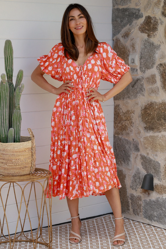 Woman wearing an orange Chrysta V-Neck midi dress with white floral print and puff sleeves standing indoors