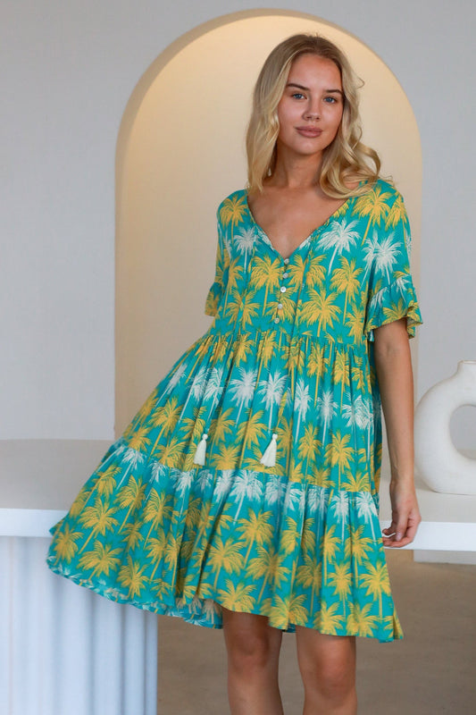 Woman wearing a colorful Clara Mini Dress with yellow and white palm tree print on turquoise background