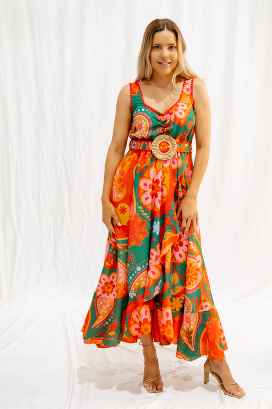 Woman wearing a colorful maxi dress with floral print, rattan belt, and gathered bust standing against a white backdrop