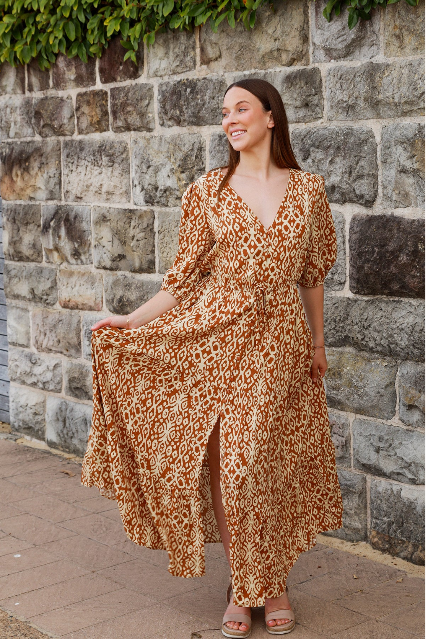 Woman wearing a patterned Cora Maxi Dress by Solara Boheme standing by a stone wall with a flowing skirt and smiling.