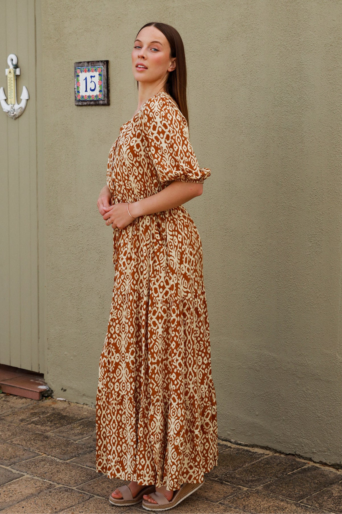 Woman wearing Solara Boheme Cora Maxi Dress with unique print standing outdoors by a wall and door.