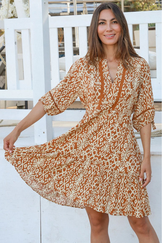 Woman wearing a brown and white patterned Cora Mini Dress with three-quarter sleeves, standing outdoors by a white fence