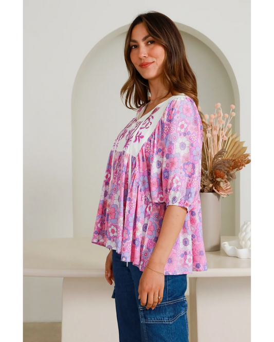 Woman wearing a loose-fitting Eliana Blouse with pink floral print and embroidered white yoke, paired with blue jeans