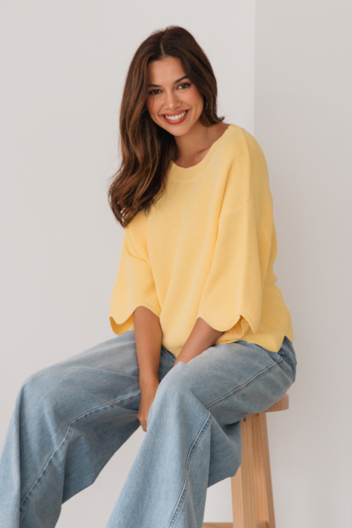 Woman wearing a yellow sweater and blue jeans sitting on a wooden stool against a white wall.