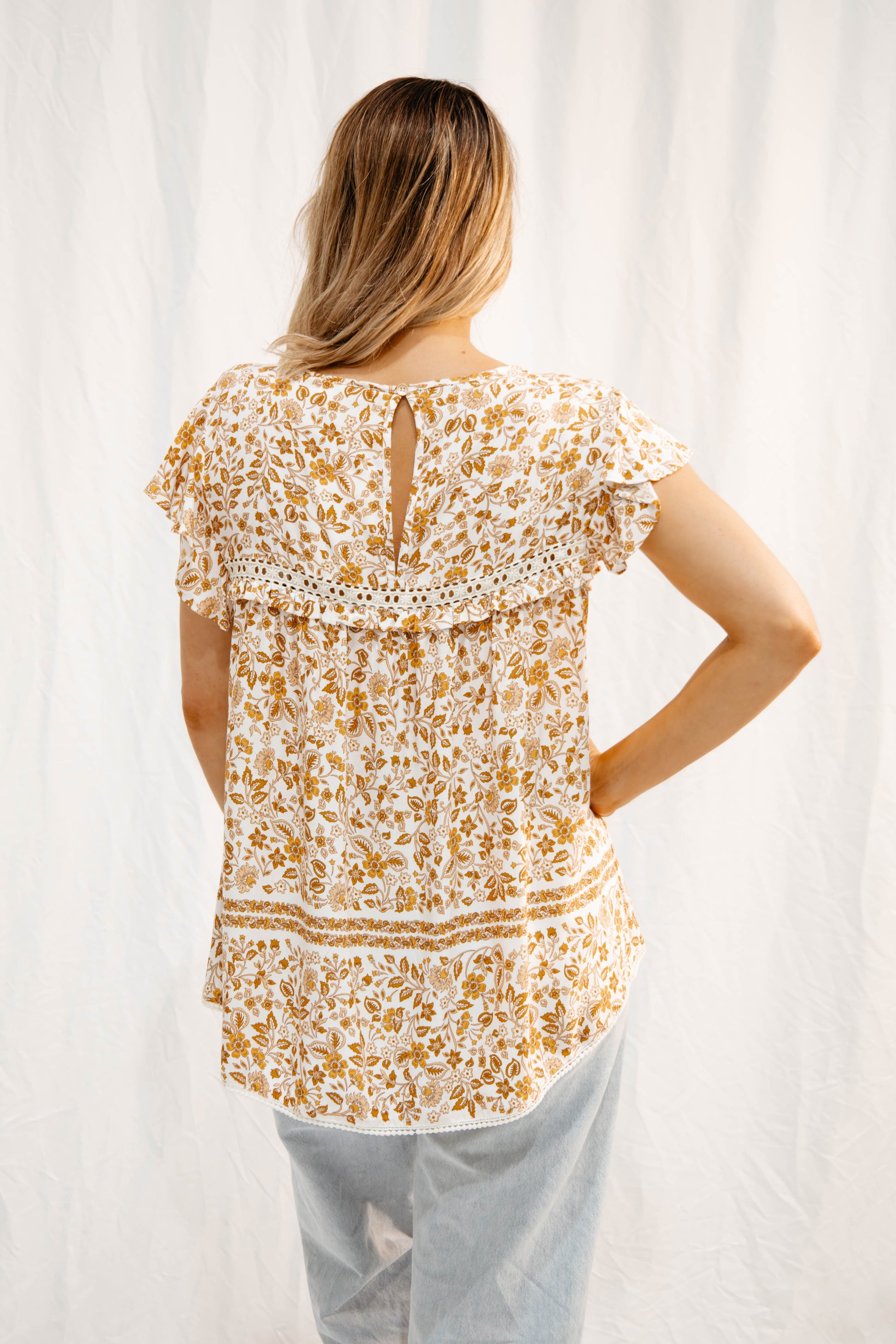 Back view of Solara Boheme Meadow Flutter Blouse with golden floral print and flutter sleeves on model