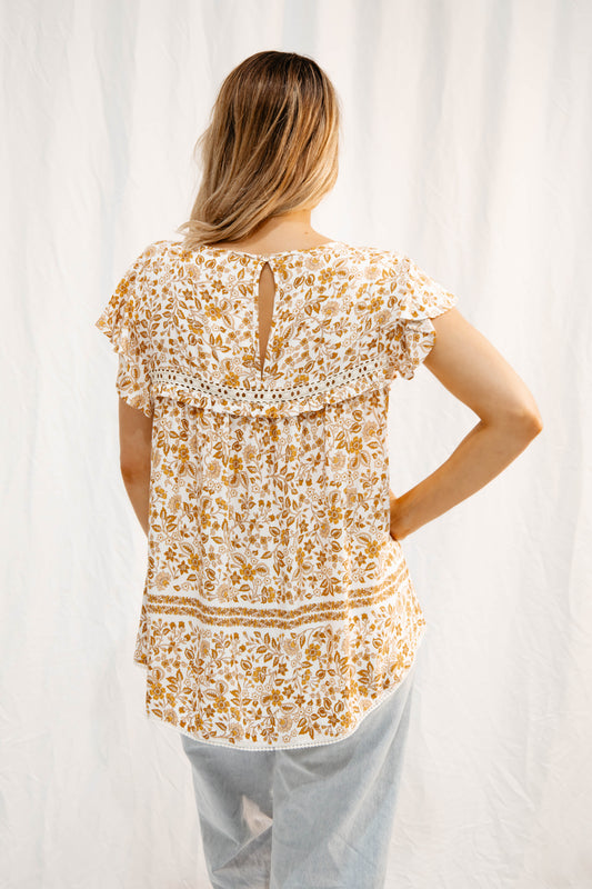 Back view of Solara Boheme Meadow Flutter Blouse with golden floral print and flutter sleeves on model