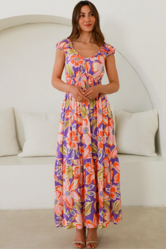 Woman wearing a colorful floral Mira Maxi Dress from My Store standing indoors with hands clasped and neutral background.