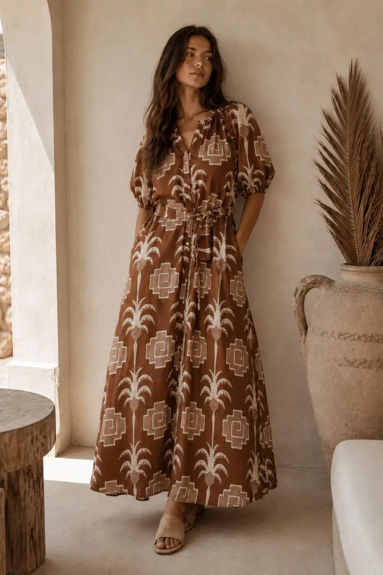 A woman wearing a brown and beige bohemian maxi dress with a geometric palm tree pattern, standing in a warm-toned interior next to a large decorative vase.