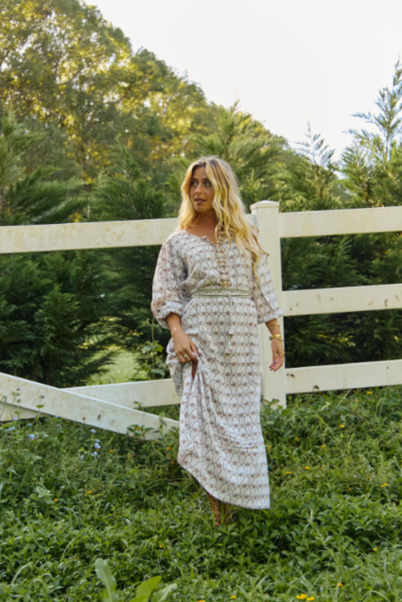 Woman wearing a Palm Springs Maxi Dress by Solara Boheme standing in a green field near a white wooden fence