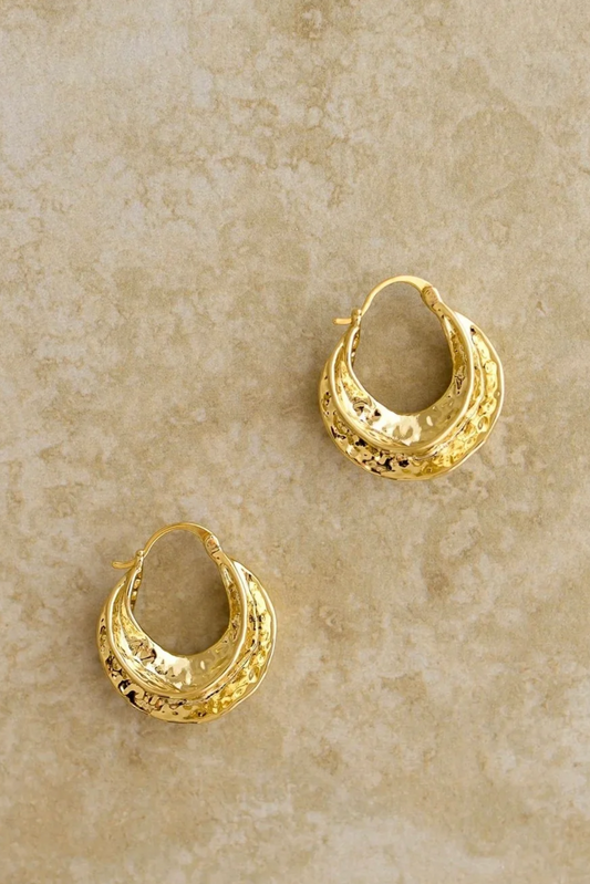 Reeve Earrings by My Store Gold plated hoop earrings with a sleek hammered design and luxurious shine on beige background.