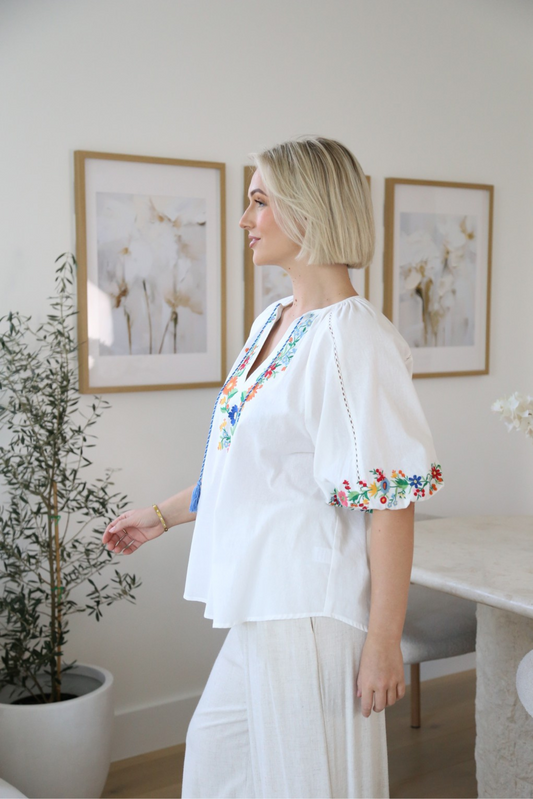 Woman wearing Retro Garden Blouse by Solara Boheme featuring colorful floral embroidery and loose sleeves in a bright room