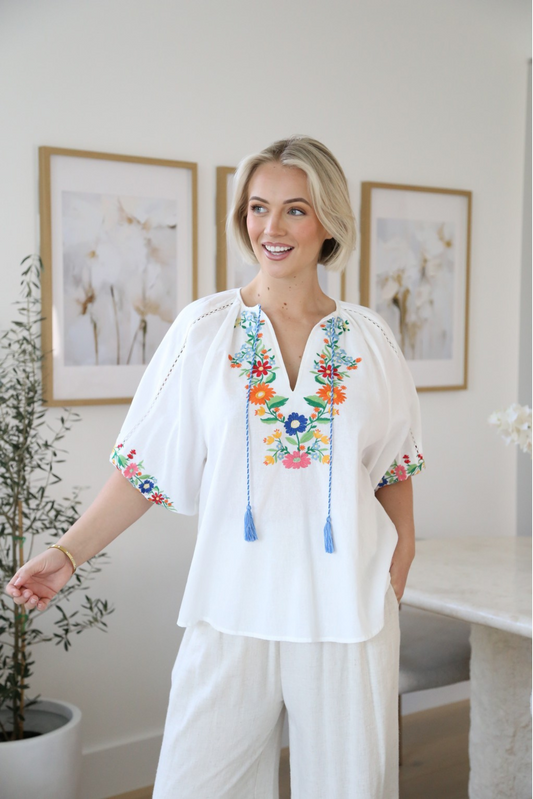 Woman wearing a Solara Boheme Retro Garden Blouse with colorful floral embroidery and tasseled ties in a bright room