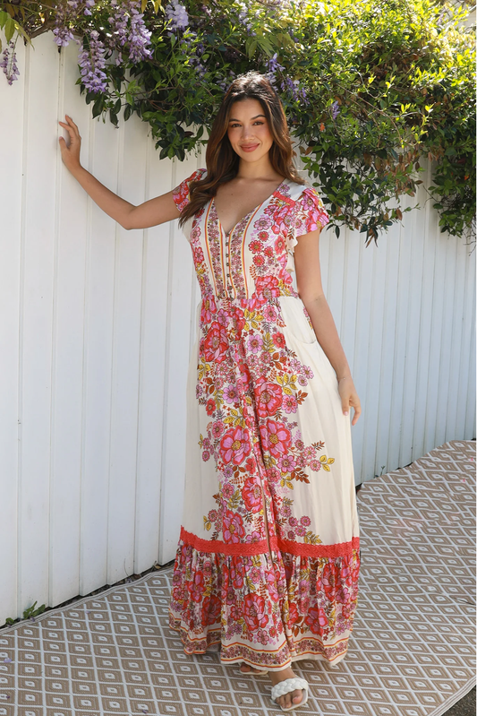 Woman wearing Solara Boheme Rosalie Maxi Dress with vibrant floral print standing by white fence under flowers