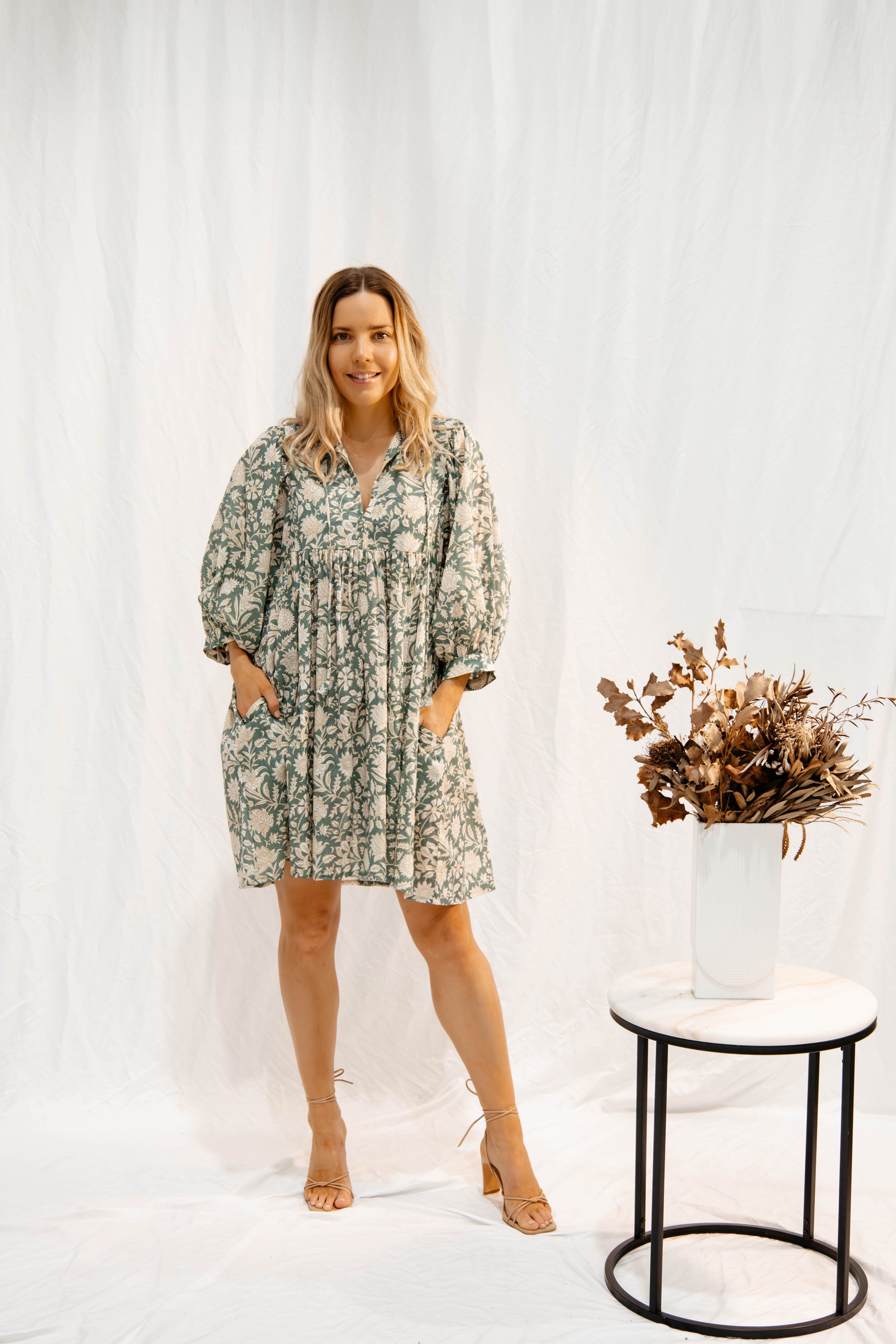 Woman wearing Solara Boheme’s Sara Mini Dress in soft olive green with floral hand block print and pockets, standing by table with dried flowers