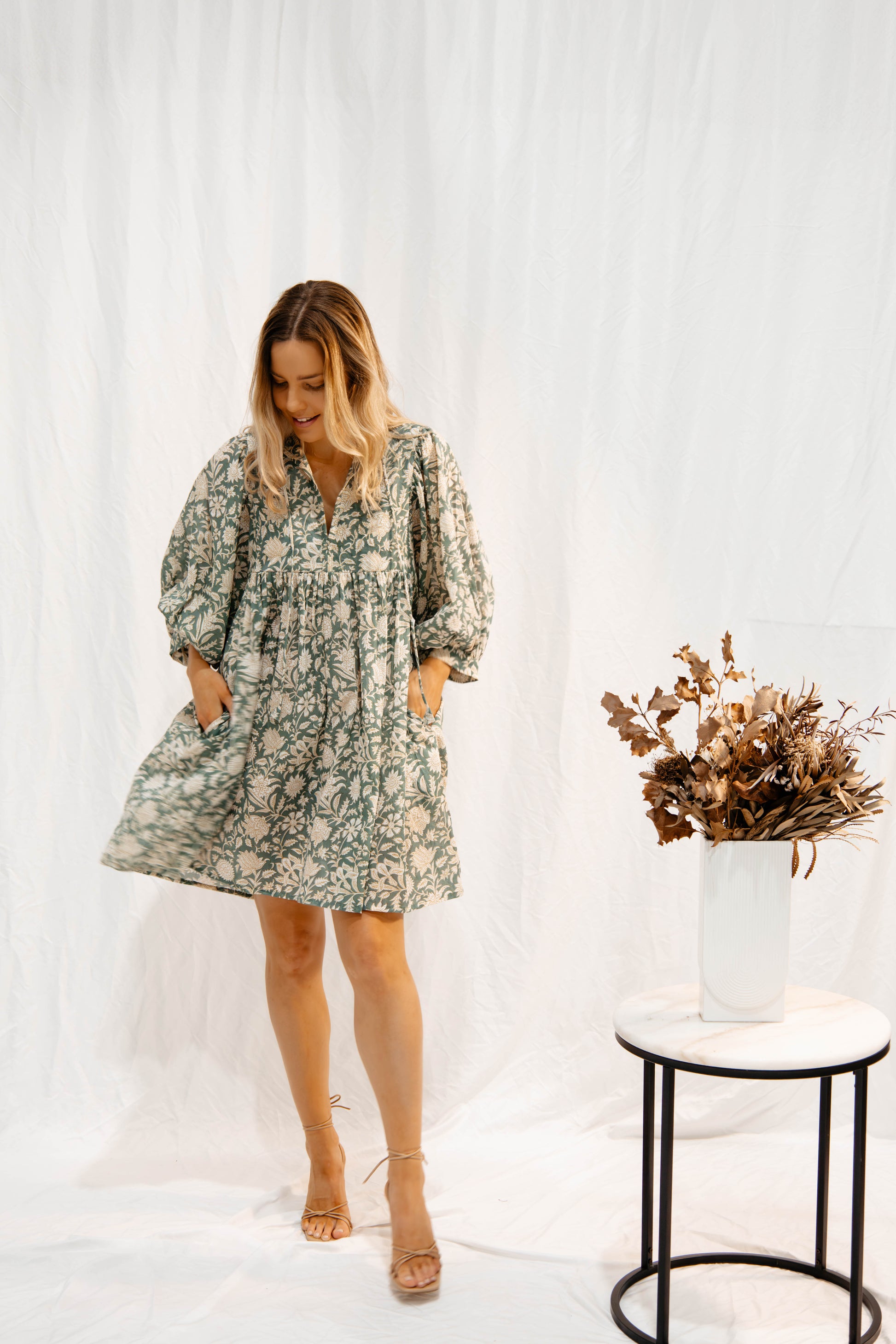 Woman wearing a Solara Boheme Sara Mini Dress in soft olive green with floral hand block print and pockets