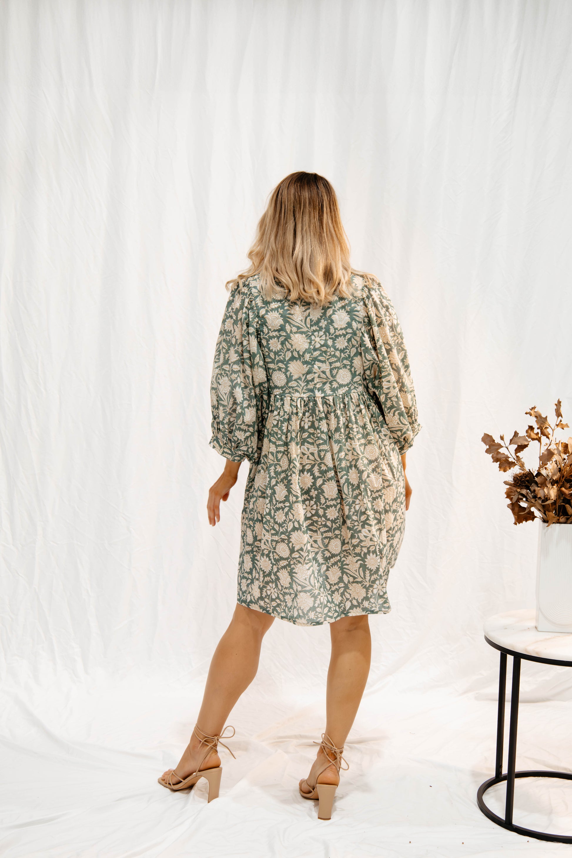 Back view of woman wearing Solara Boheme Sara Mini Dress with floral pattern and flowy sleeves standing by table