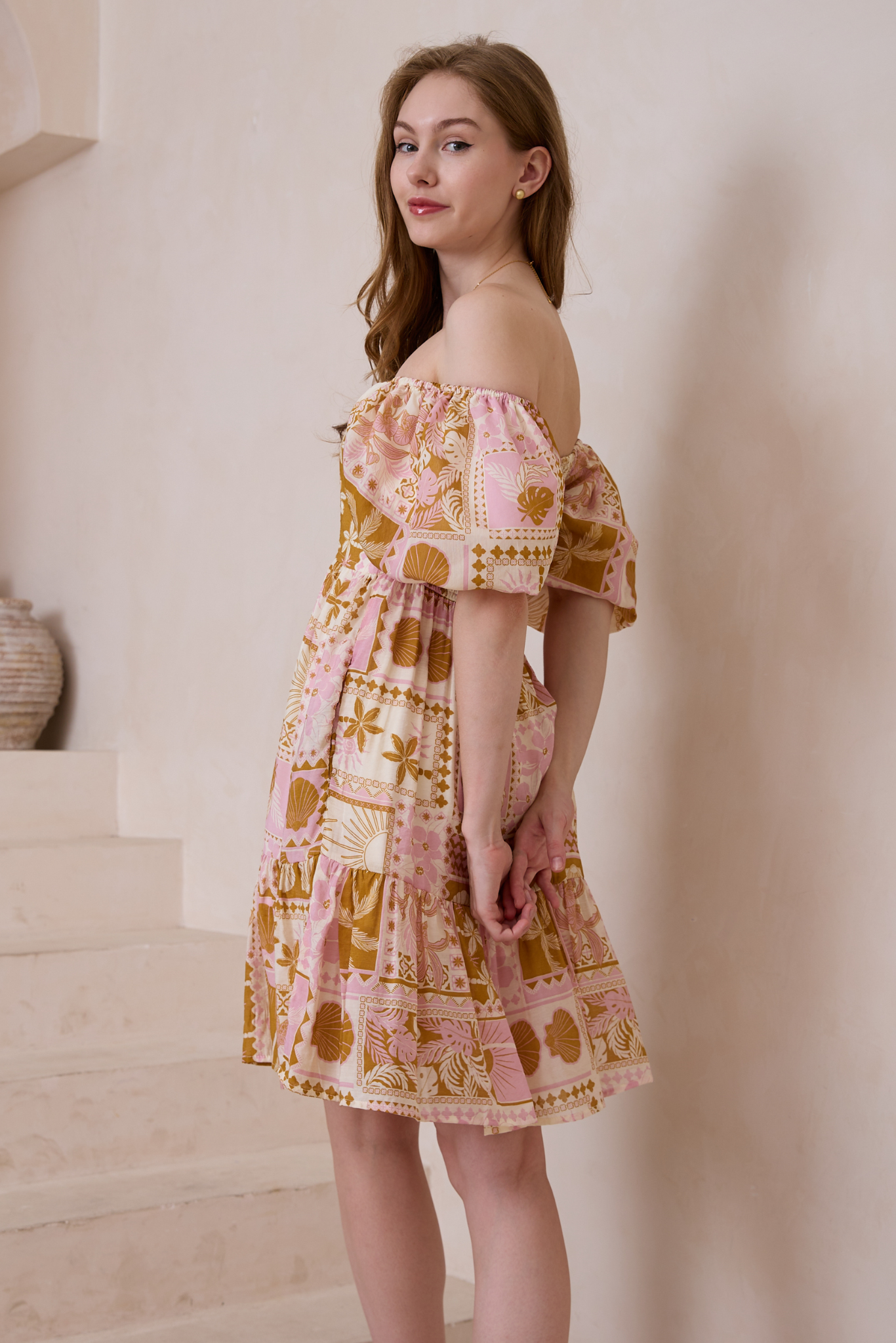 Woman wearing a patterned off-shoulder Shelley Mini Dress by Solara Boheme with shirred back and side seam pockets.