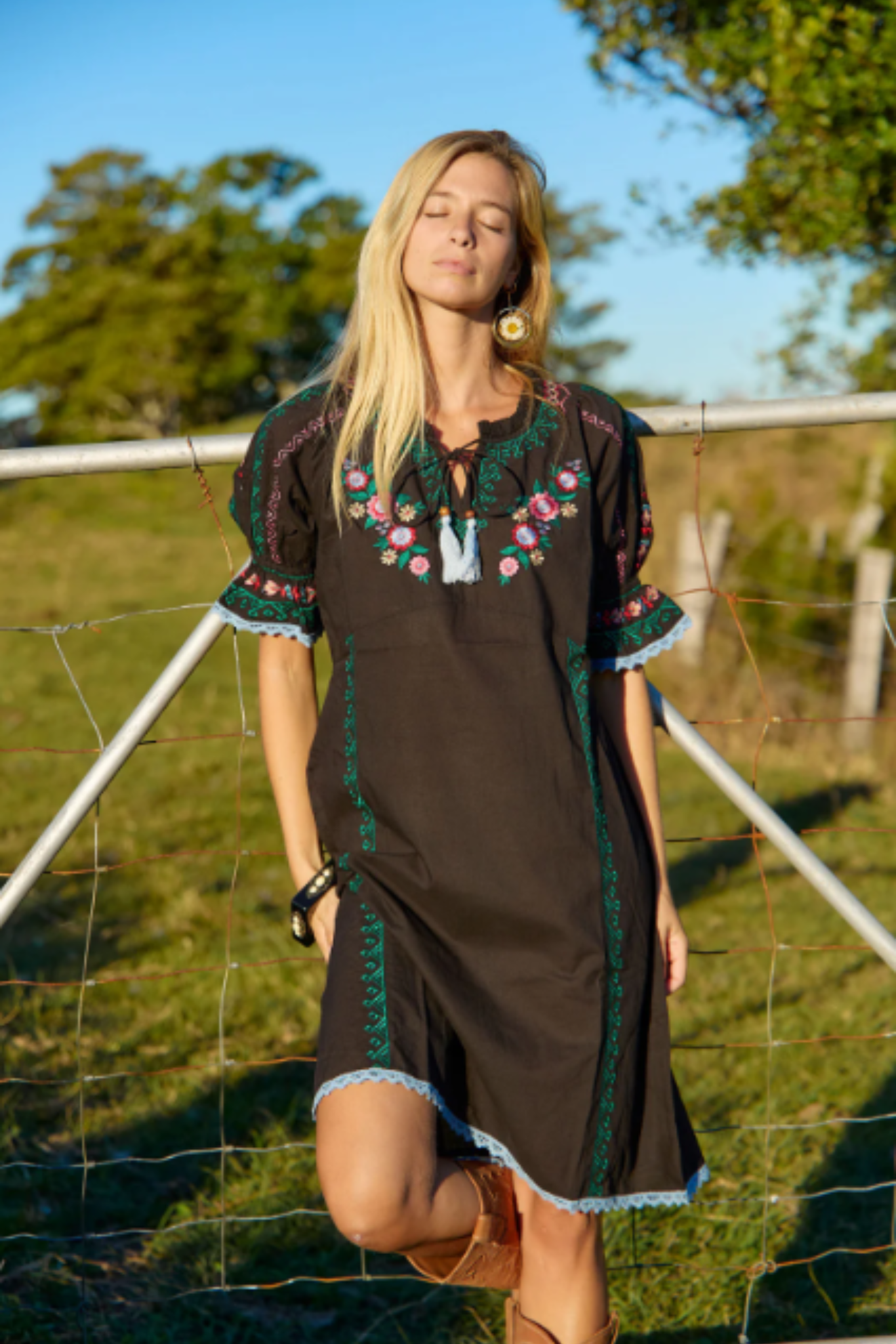 Woman wearing Solara Boheme Spell Mini Dress with floral embroidery standing outdoors by a fence in sunlight