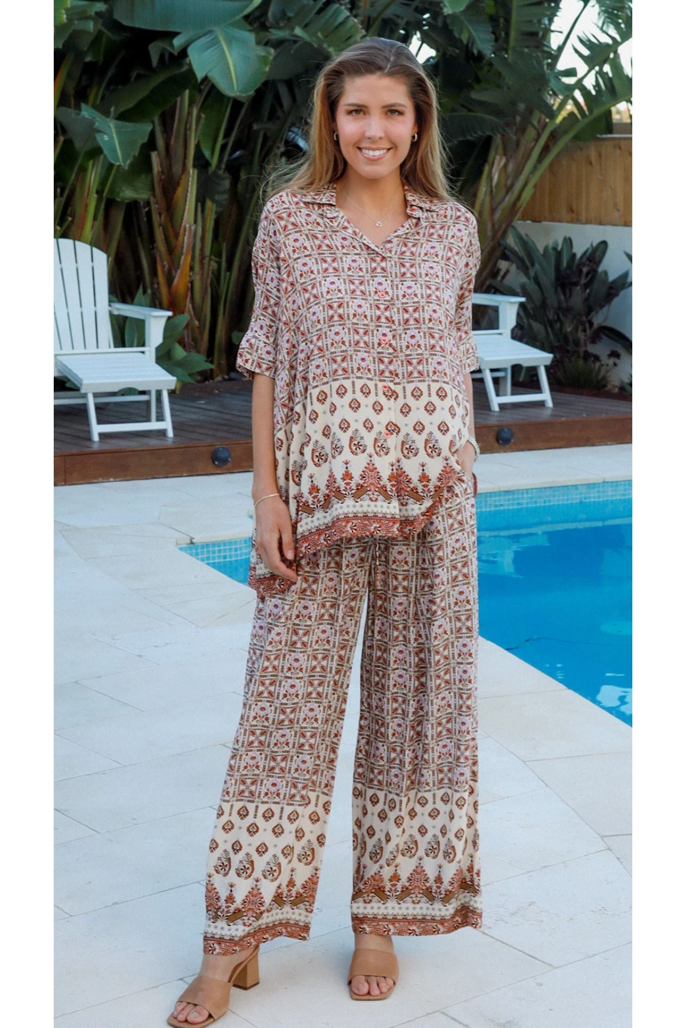 Woman wearing patterned Tori Pants by My Store standing near pool with tropical plants in background