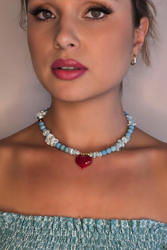 A woman wearing a blue off-the-shoulder, patterned top and a beaded choker necklace made of turquoise and white stones with a small red heart pendant in the center. She has light makeup, pink lipstick, long eyelashes, and her hair pulled back.