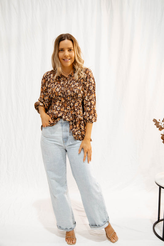 Woman wearing Solara Boheme Wildflower Whisper Blouse with brown floral print and light-wash jeans in relaxed pose
